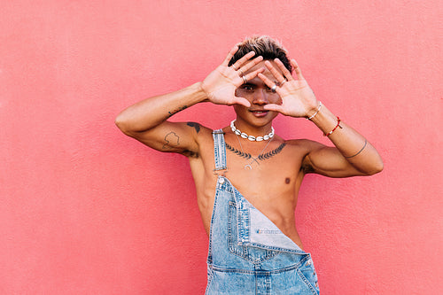 Young handsome guy hiding his face with palms while looking at camera. Stylish man in overalls standing outdoors at a pink wall.