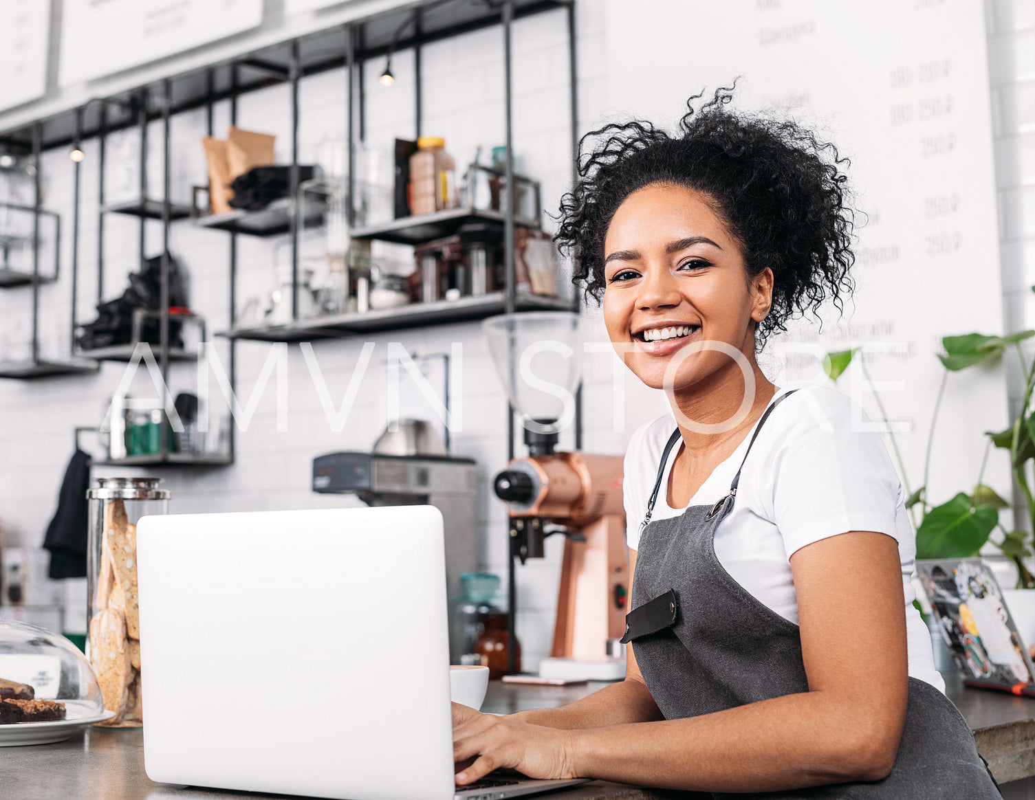 Smiling coffee shop owner with curly hair looking at the camera. Woman in an apron with a laptop at counter.