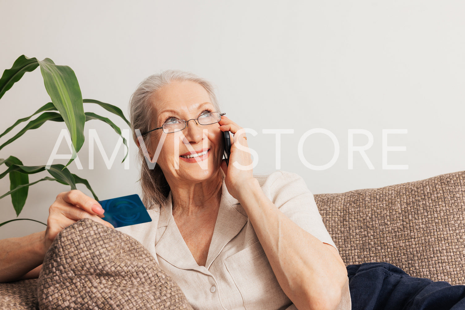 Smiling senior woman ordering through a cell phone. Aged female in eyeglasses holding a credit card while lying on a sofa.