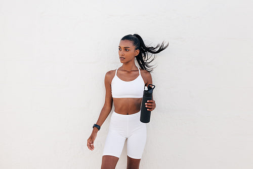 Portrait of a slim sportswoman with a shaker at a white wall. Female in white fitness attire holding a bottle.