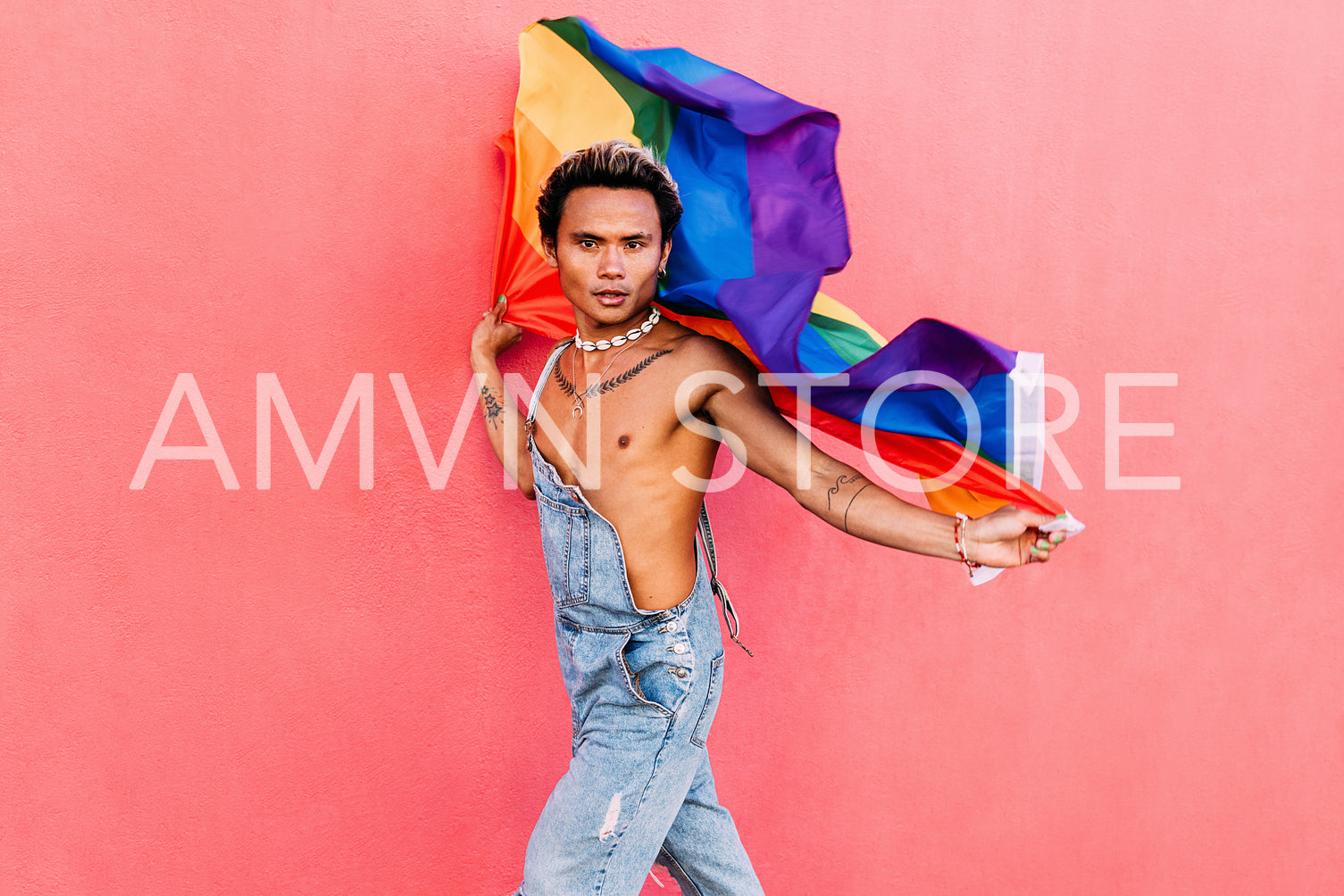 Young stylish man holding a waving LGBT flag against pink wall outdoors and looking at camera