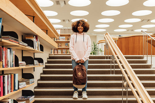 Schoolgirl with backpack standing on stairs in library. Smiling student in casuals looking at camera.