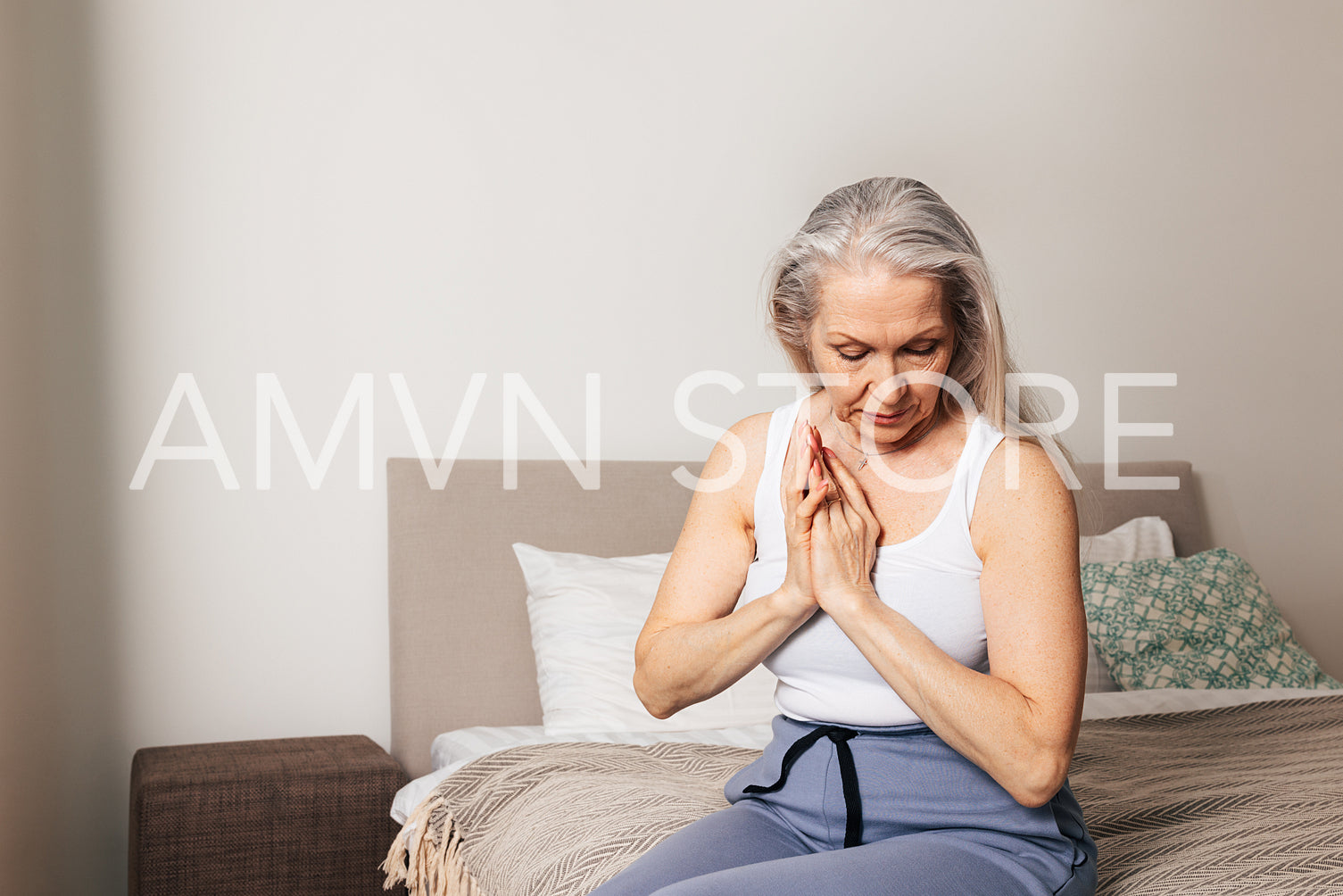 Senior woman with grey hair sitting on bed with folded hands and looking down