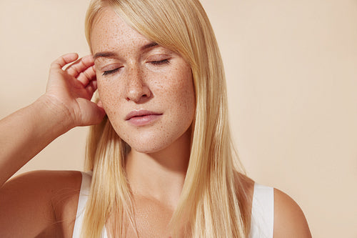 Young female with freckled skin standing with her eyes closed in studio. Blond woman adjusting hair while posing against pastel backdrop.