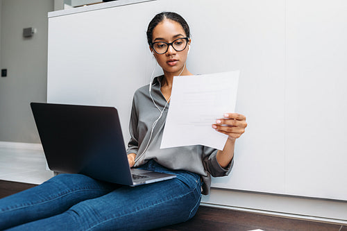 Young woman sitting on floor at kitchen counter with document in hand and using laptop