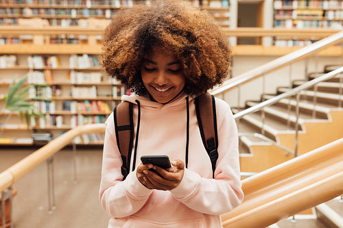 Smiling girl holding smartphone in library against bookshelf