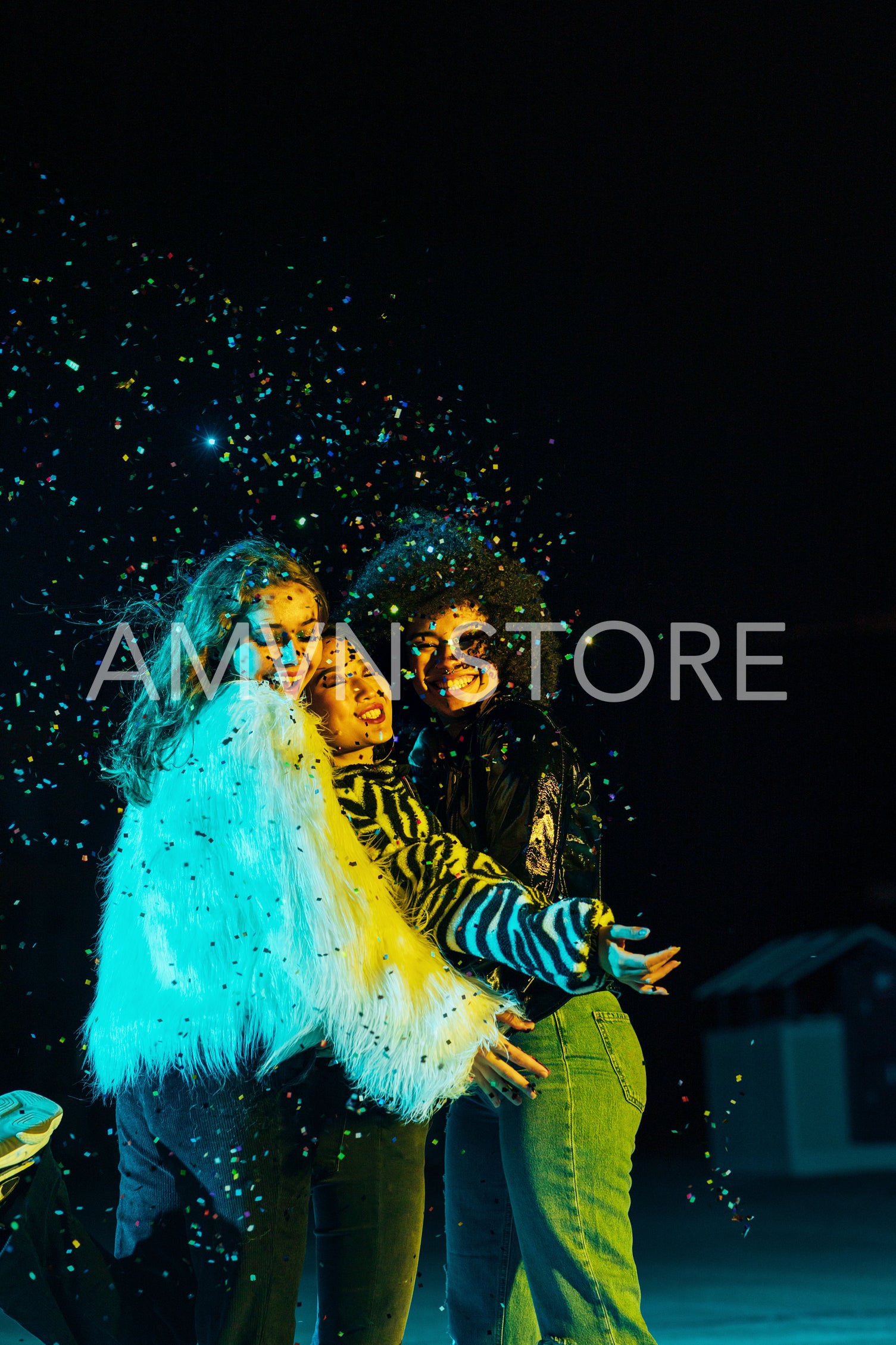 Three happy female friends embracing each other while standing under confetti at night	