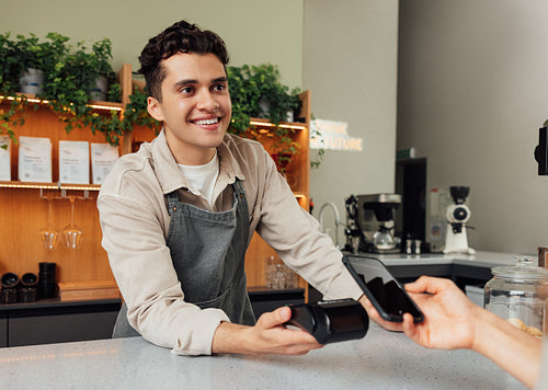 Smiling male barista holding pos terminal looking at the customer. Bartender receive contactless payment.