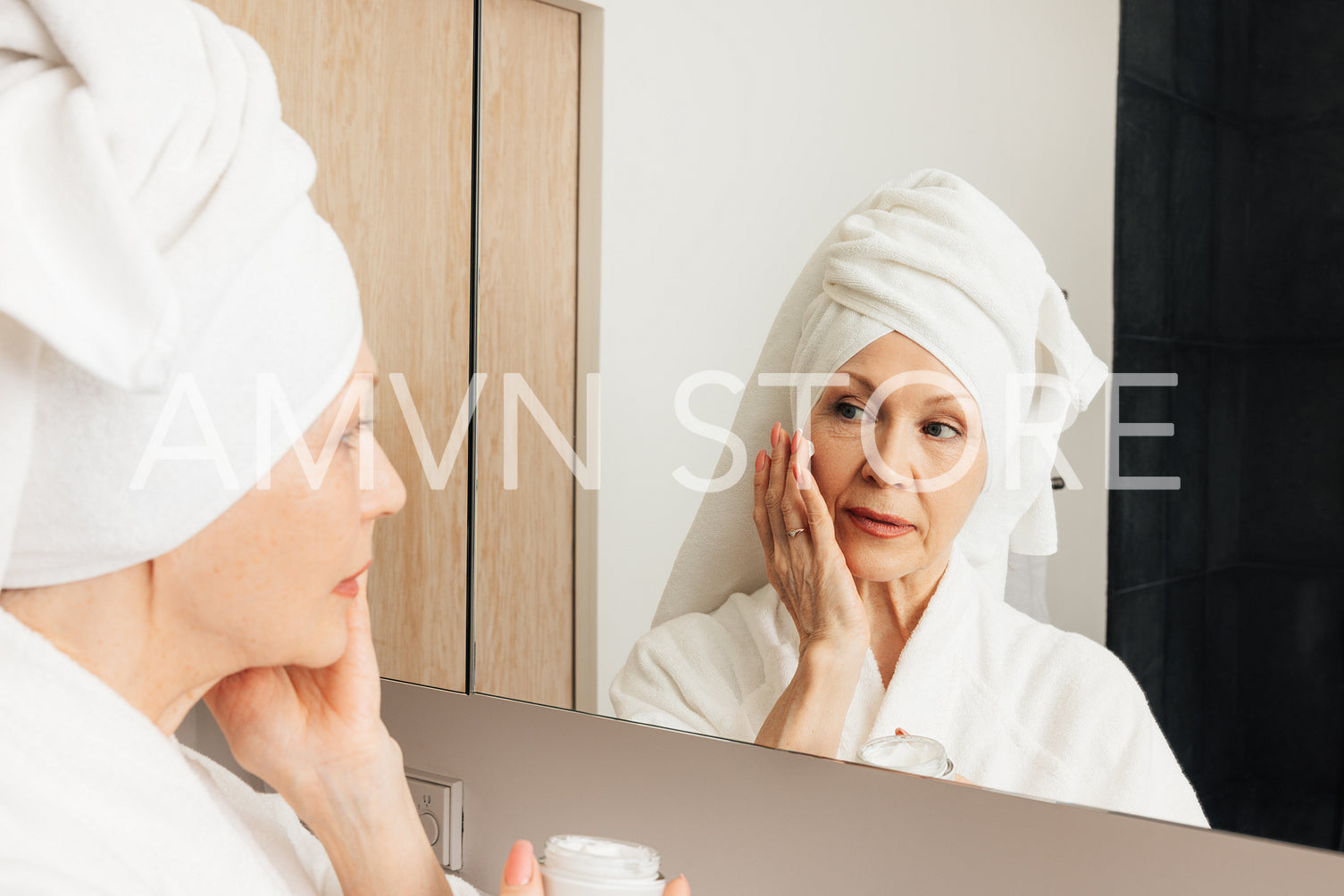 Senior woman with wrapped towel on her head applying cream on a face. Aged female doing morning routine in front of a bathroom mirror.