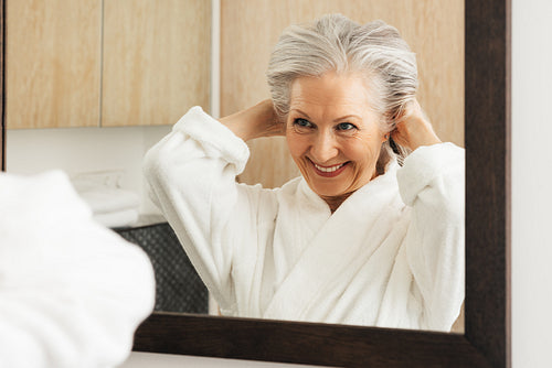 Smiling aged woman making funny hairstyle while looking at a mirror in bathroom. Senior female enjoying morning routine in the bathroom.