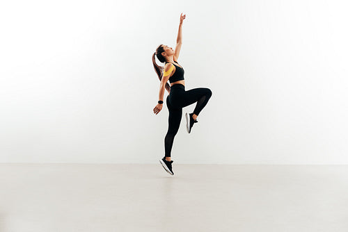 Woman jumping in the air against white wall raising one hand up. Young female in sports clothes warming up.