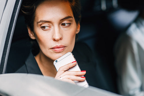 Thoughtful woman holding a cell phone while sitting on a backseat of a taxi