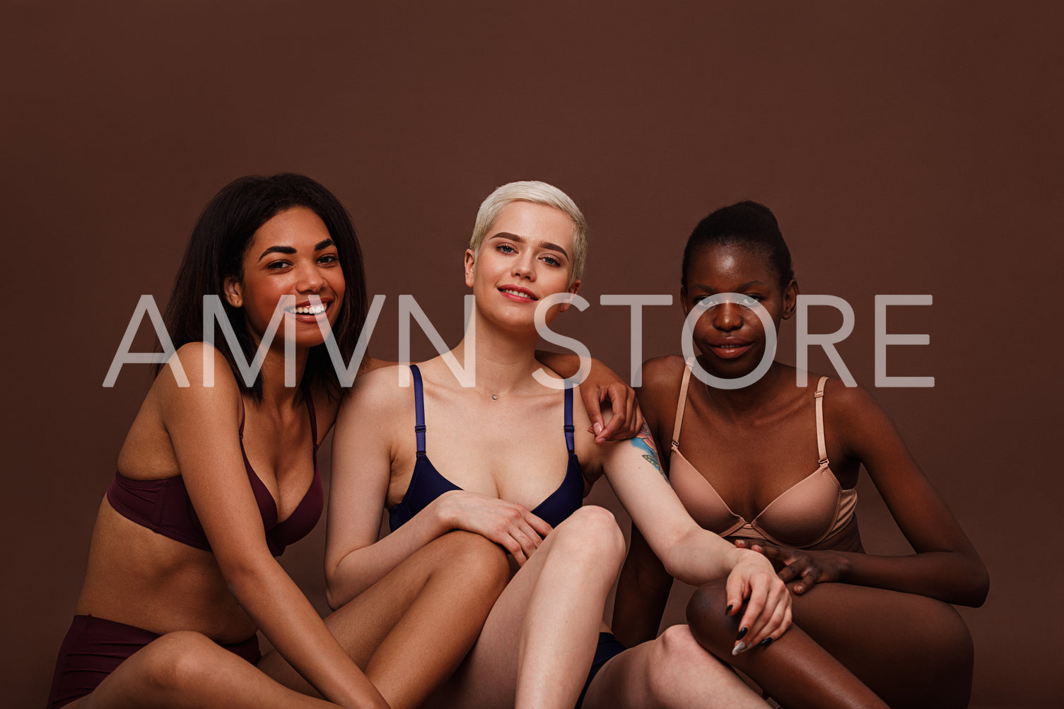 Three women with different skin color sitting together against brown background and smiling