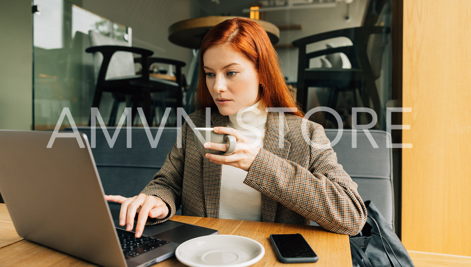 Woman working as a digital nomad in a cafe. Female entrepreneur with ginger hair holding a cup typing on a laptop