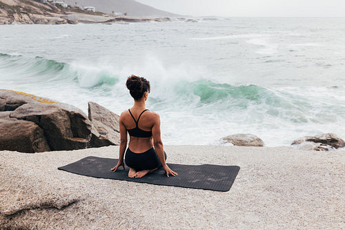 Rear view of young woman sitting in Vajrasana pose on mat outdoors. Woman meditating while looking at ocean.