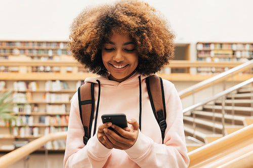 Close up of smiling schoolgirl standing in library and using smartphone