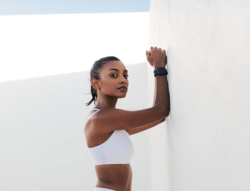 Confident slim female relaxing at the wall. Young fitness influencer in sports bra looking at camera.