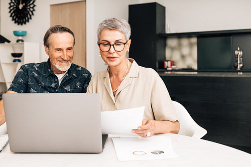 Senior retired couple at a table in dining room using a laptop computer. Two mature people looking at paperwork and discussing their finances.