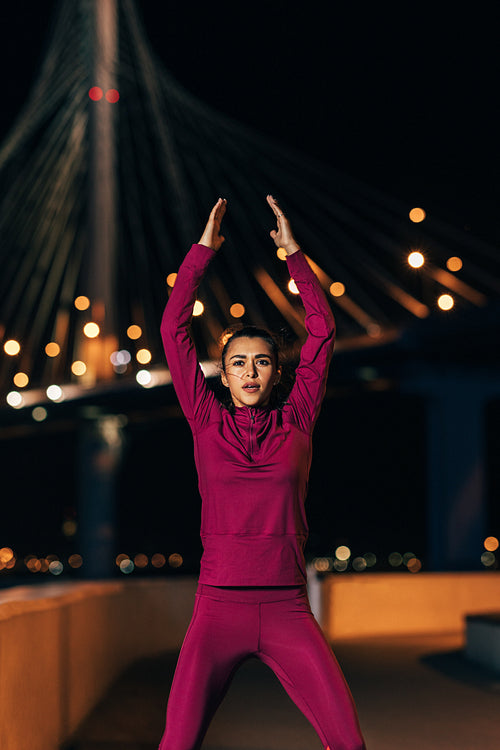 Female athlete in front of bridge at night. Woman wearing sportswear warming up body in the city in the evening.