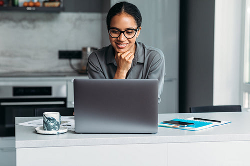 Female entrepreneur working at home office. Smiling woman standing at desk in kitchen and looking on a laptop screen.