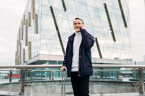 Mature man standing in front of office building with suitcase and talking on cell phone