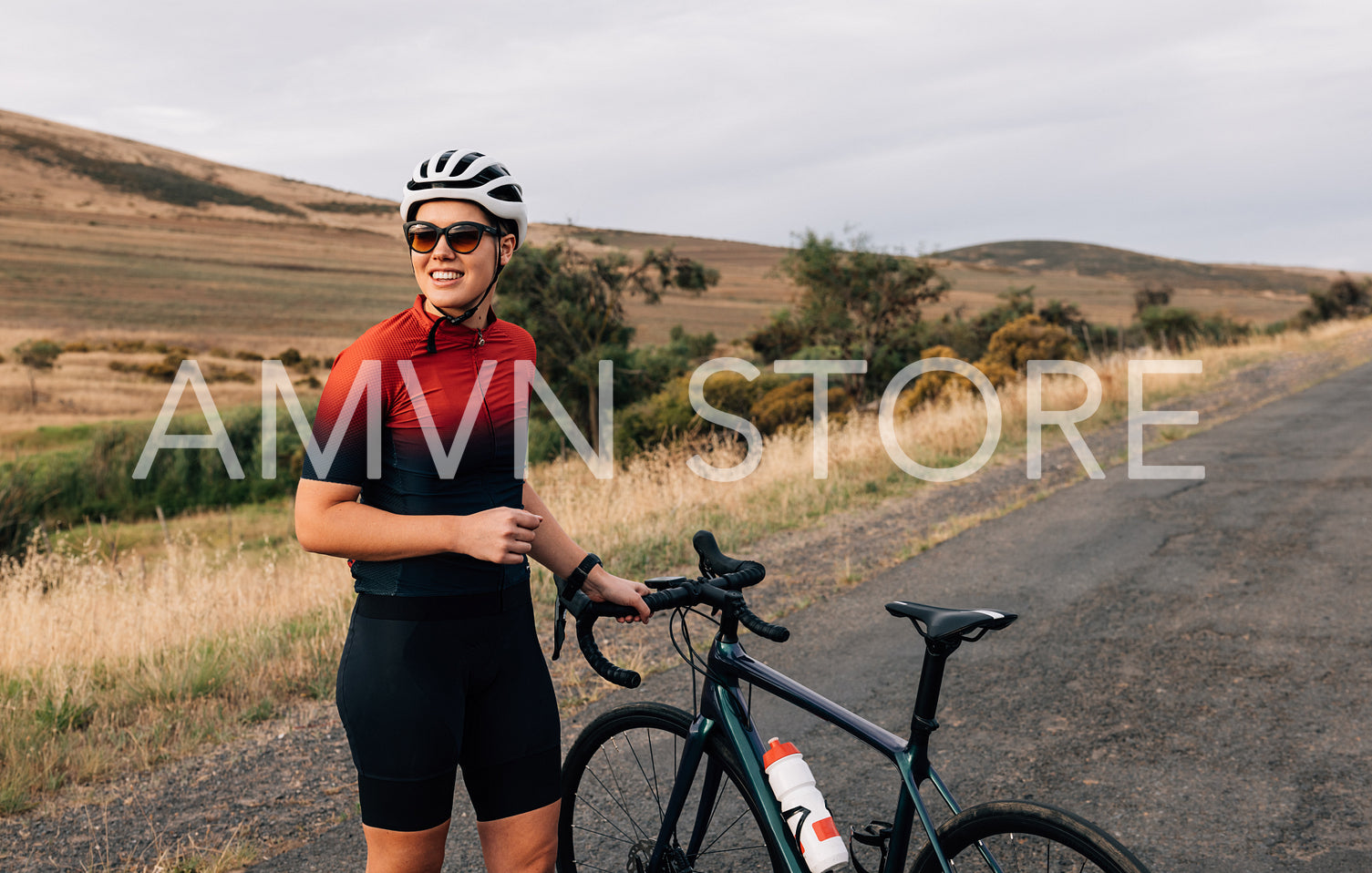 Smiling woman cyclist wearing sunglasses and helmet taking a break during ride standing on a road