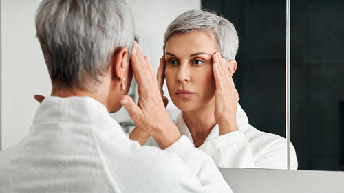 Mature woman in a bathroom in front of a mirror touching head with hands