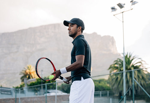 Fit man in sportswear and cap with a racket standing on tennis court. Tennis player looking away while standing on a hard court.