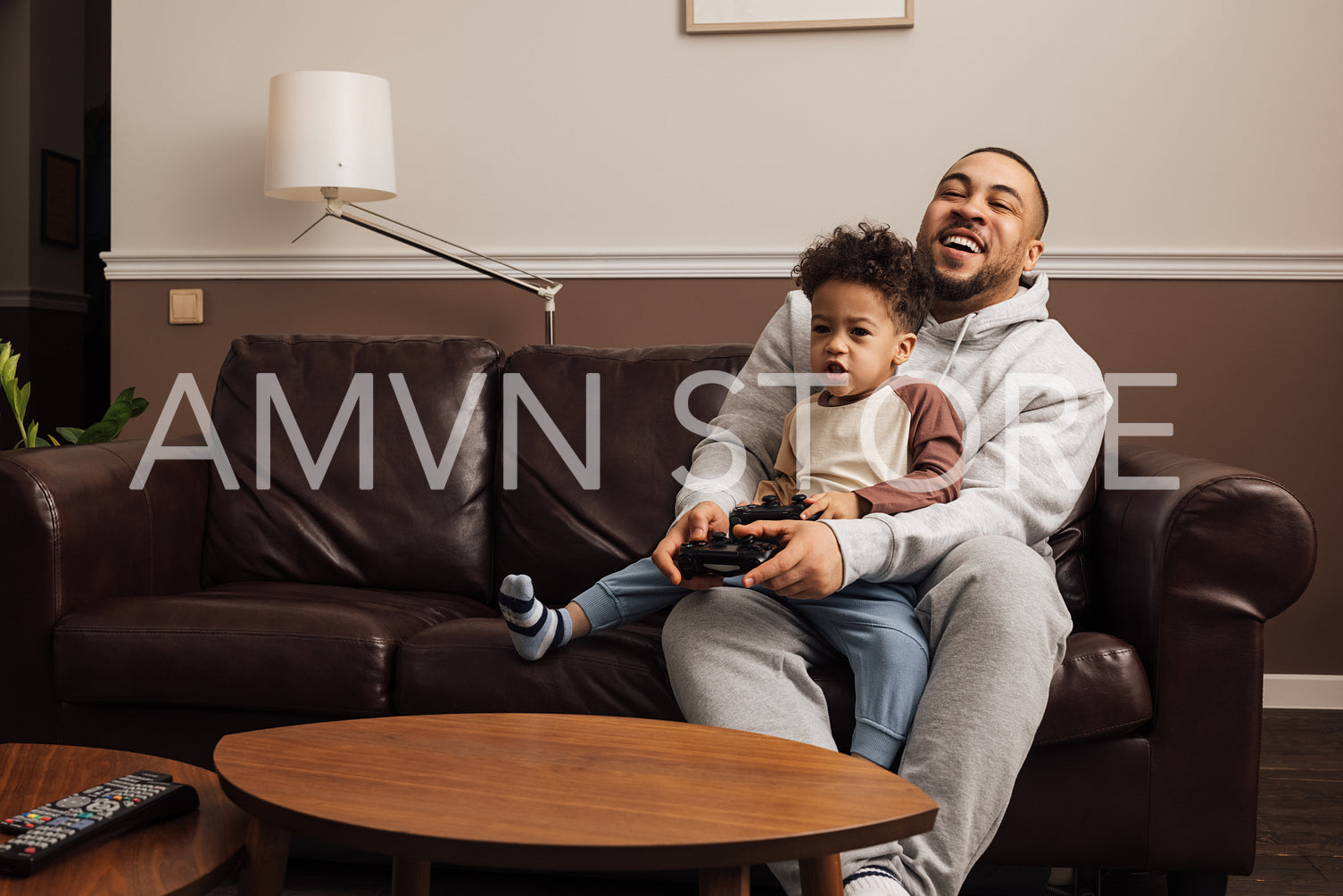 Father and son playing a video game together at home. Young man and little boy sitting on sofa holding joysticks and having fun.