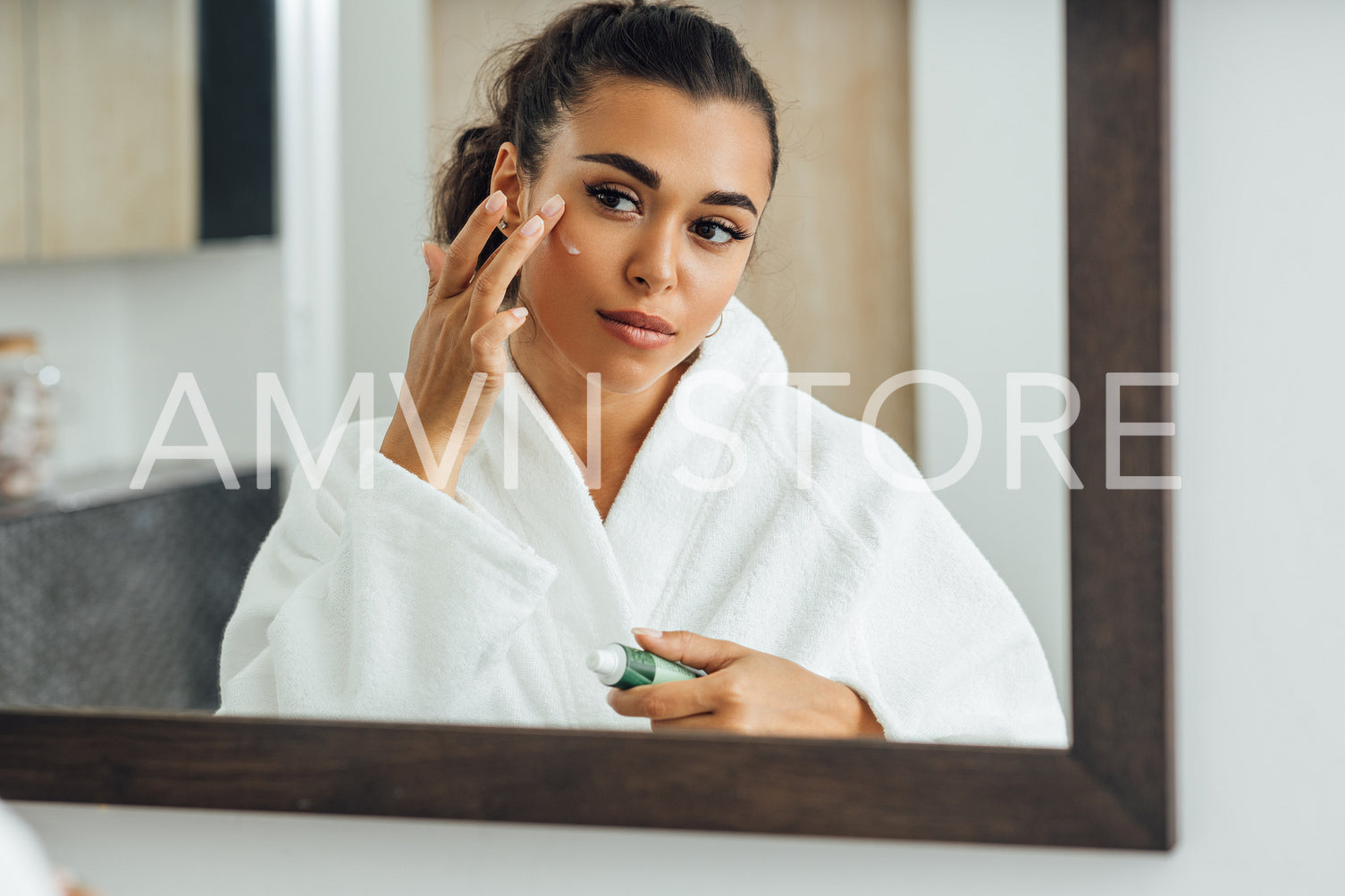 Young woman applying face cream in front of a mirror. Middle east female using a moisturizer after bath.	