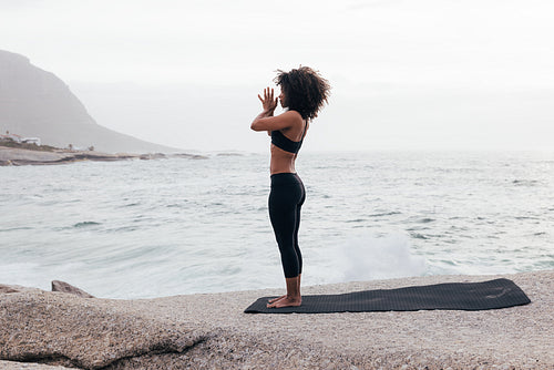 Full length of slim female standing with joined hands and meditating by ocean at evening