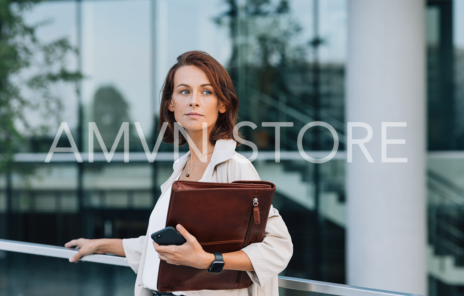 Confident female in coat with leather folder looking away while standing at railing against business building