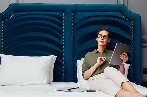 Young woman holding a digital tablet and looking away while lying on a bed in a luxury hotel room