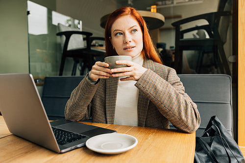 Woman with ginger hair holding a cup and looking at the window at a cafe. Businesswoman enjoying her coffee.