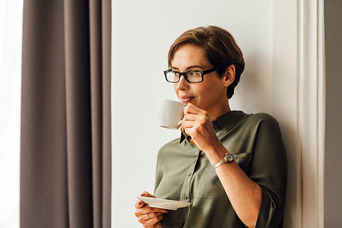 Mid adult female in formal clothes having coffee in hotel room and looking away. Business woman standing at wall in apartment and drinking coffee.