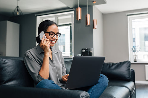 Smiling woman sitting on a sofa with laptop on her legs and making a phone call