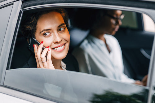 Smiling woman talking on a cell phone in a taxi. Young entrepreneur sitting on a backseat with a colleague.