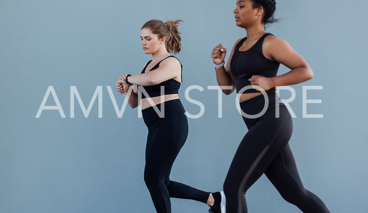 Two female friends running against a grey wall. Plus size female checking her smartwatch while jogging outdoors with her friend.