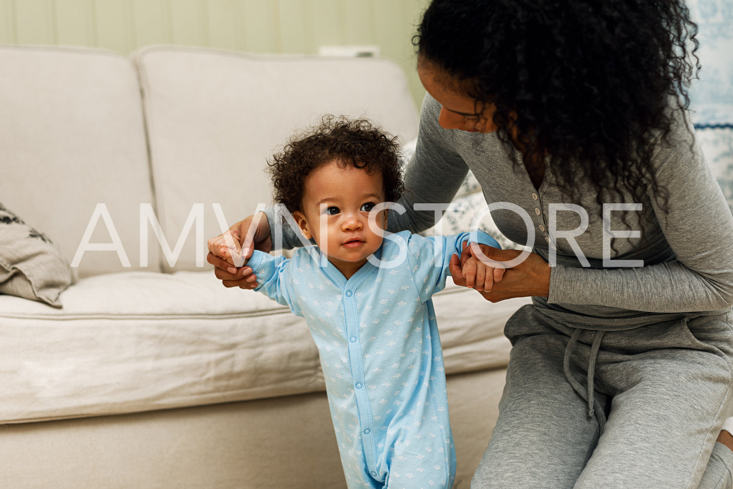 Mother sitting on the in a living room while helping her son walking	