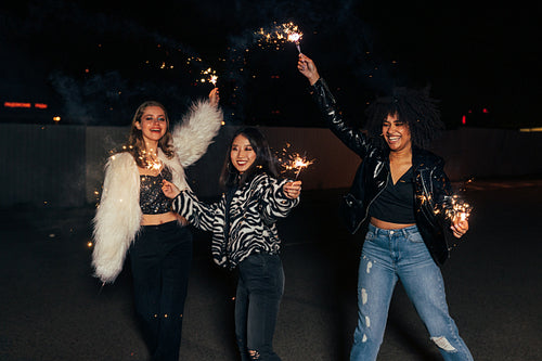 Three laughing female friends waving sparklers at night and having fun together