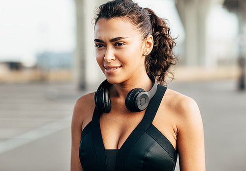 Portrait of a beautiful sportswoman with headphones on her neck. Smiling woman in sports bra standing outdoors.