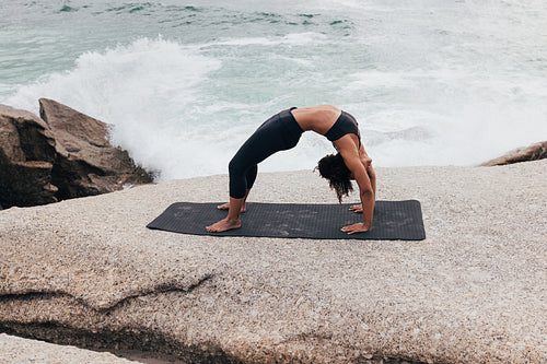 Side view of young slim female practicing Urdhva Dhanurasana. Woman in sportswear in Wheel yoga pose by ocean.