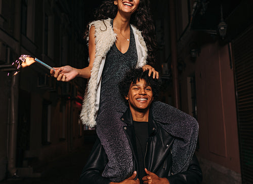 Smiling woman sitting on shoulders of her friend, holding a sparkler