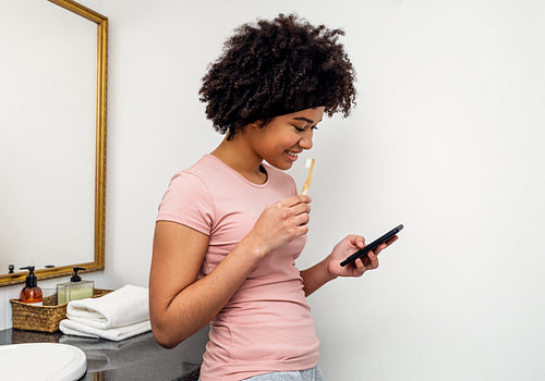 Smiling woman reading from mobile phone while brushing her teeth in bathroom