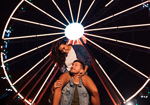 Smiling woman looking at her boyfriend while sitting on his shoulders at night in an amusement park