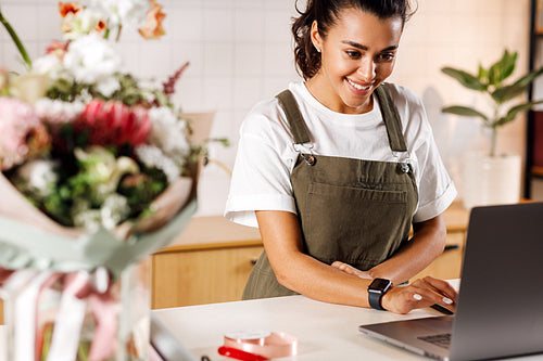 Young flower shop owner using laptop at counter. Woman florist standing at table.