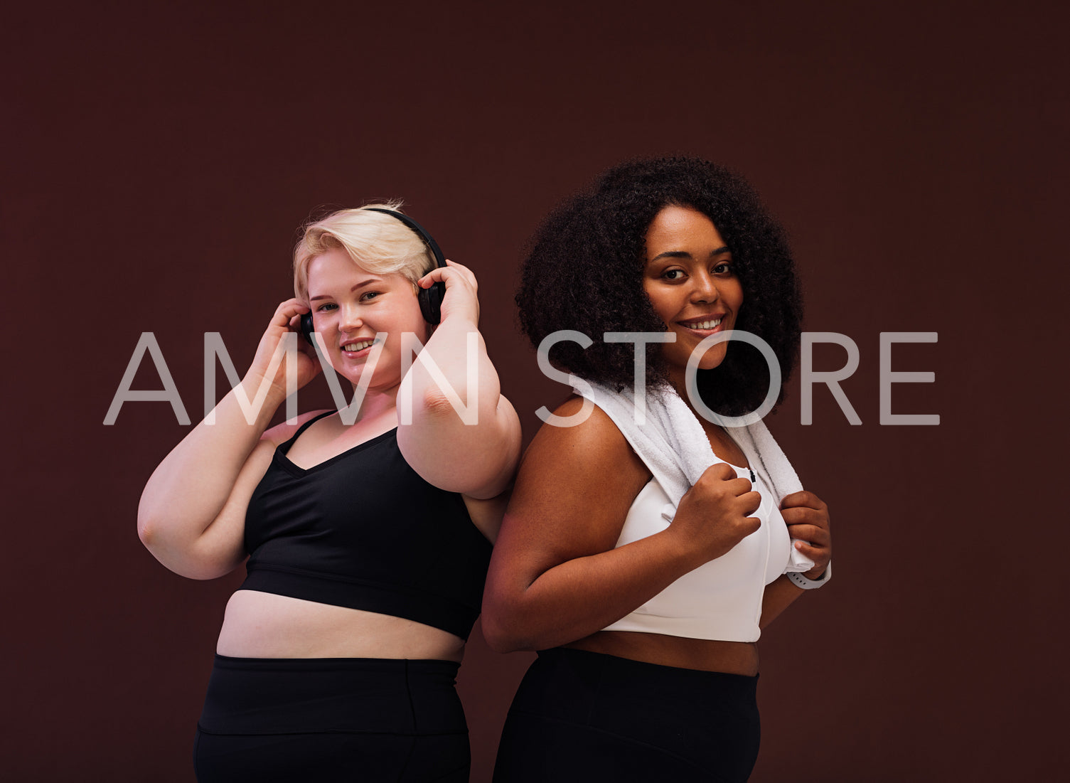 Two smiling plus size women standing together in studio. Females in sportswear posing together against brown background.