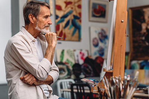 Side view of a thoughtful artist holding paintbrush while sitting in his studio. Male painter looking on canvas.