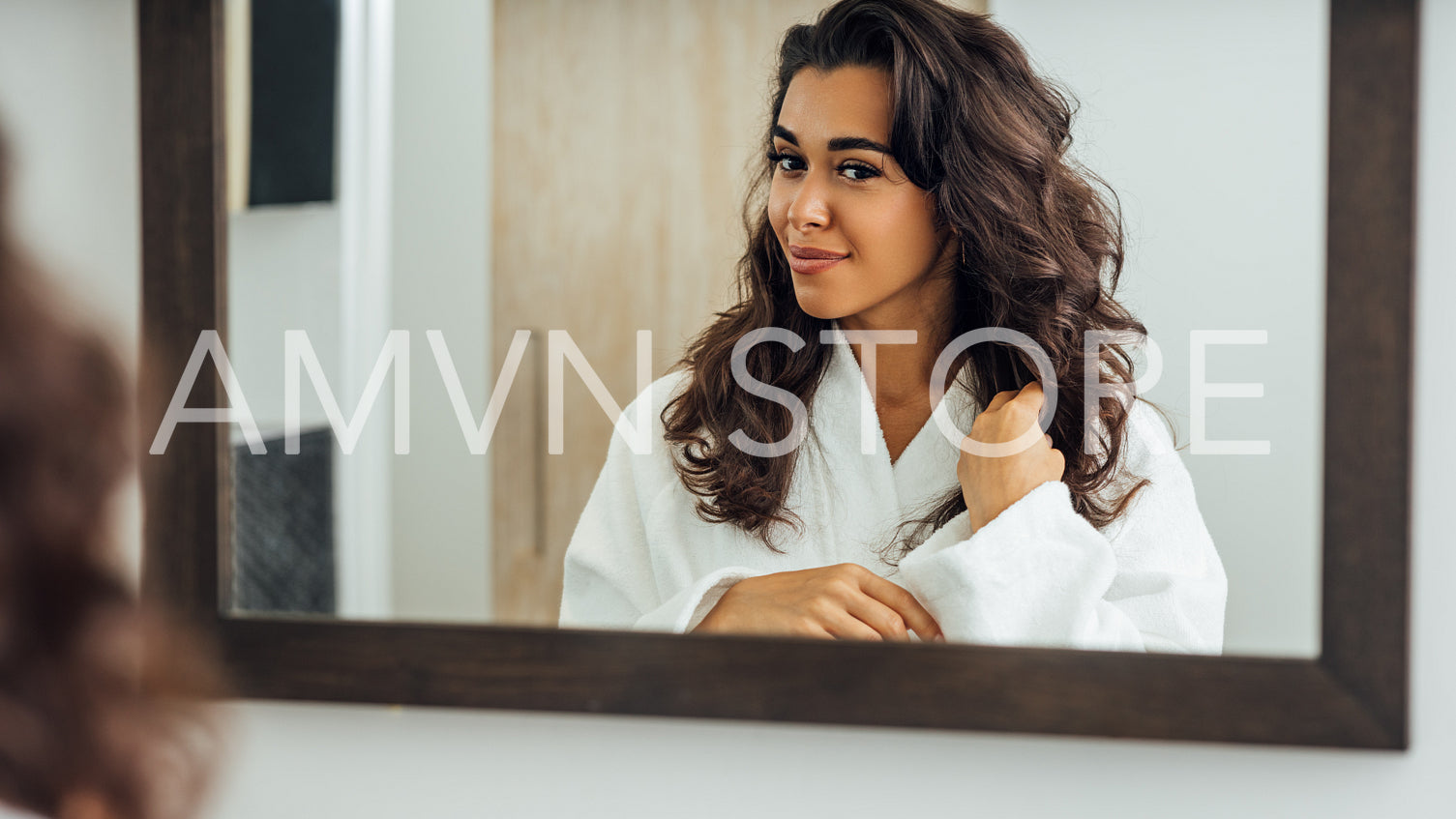 Beautiful brunette touching her hair in bathroom. Middle East woman standing in front of a mirror wearing a bathrobe.	