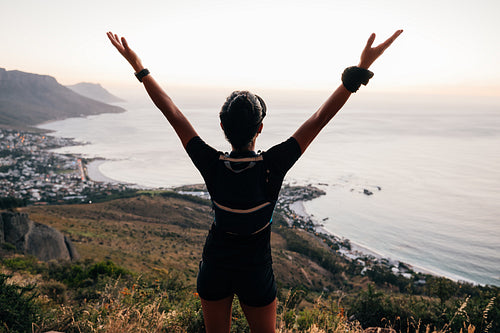 Rear view of trail runner enjoying sunset. Woman in sportswear raising hands while looking at scenic view.